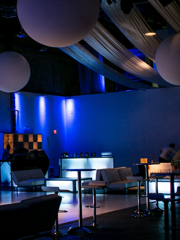 An indoor nightclub with blue lighting, modern couches, high tables, large hanging spherical and fabric ceiling decorations, a bar area with bottles, and a person working behind the bar.