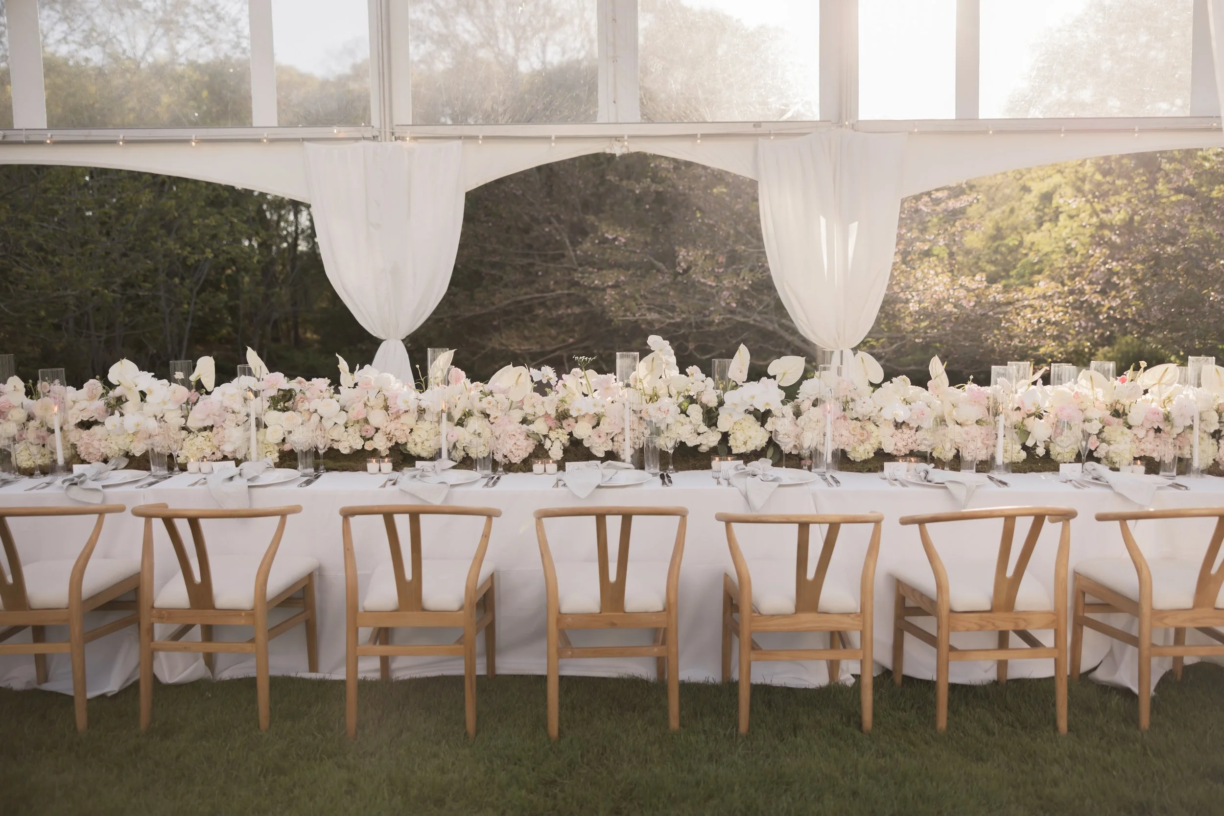 A beautifully decorated outdoor wedding reception table with a backdrop of trees and sunlight, featuring a long white table with floral arrangements, candles, and wooden chairs.