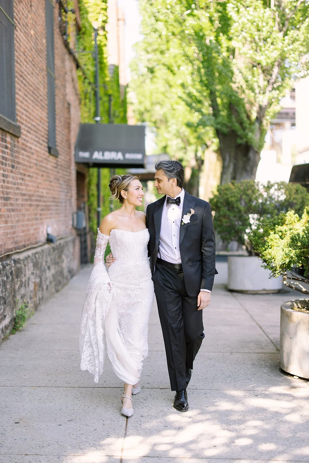 A bride and groom in wedding attire walking and smiling together outdoors on a sunny day, with trees and potted plants lining the sidewalk near a brick building.