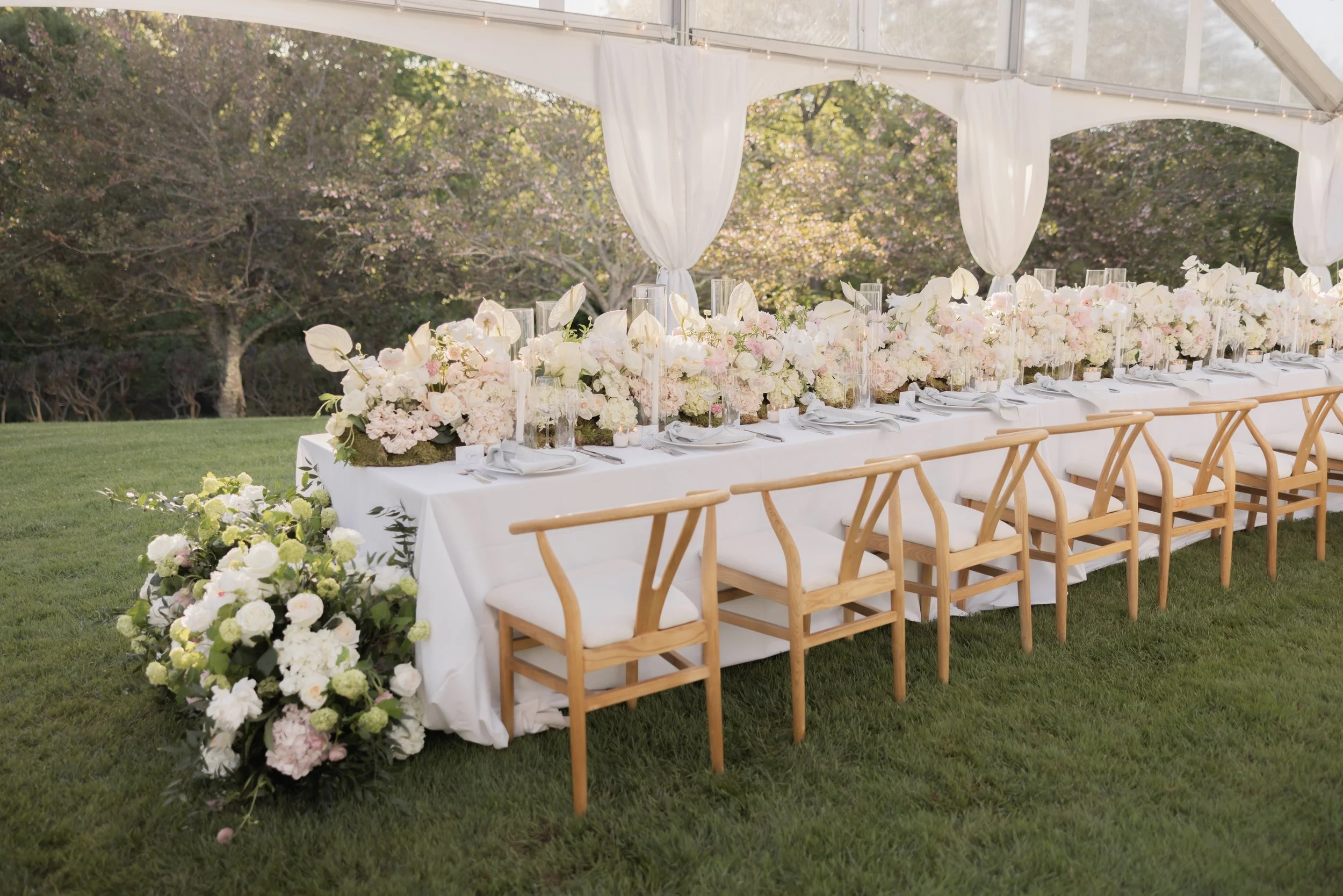 A long banquet table decorated with white and pink flowers, set up outdoors under a tent with white curtains and string lights, with wooden chairs on either side.