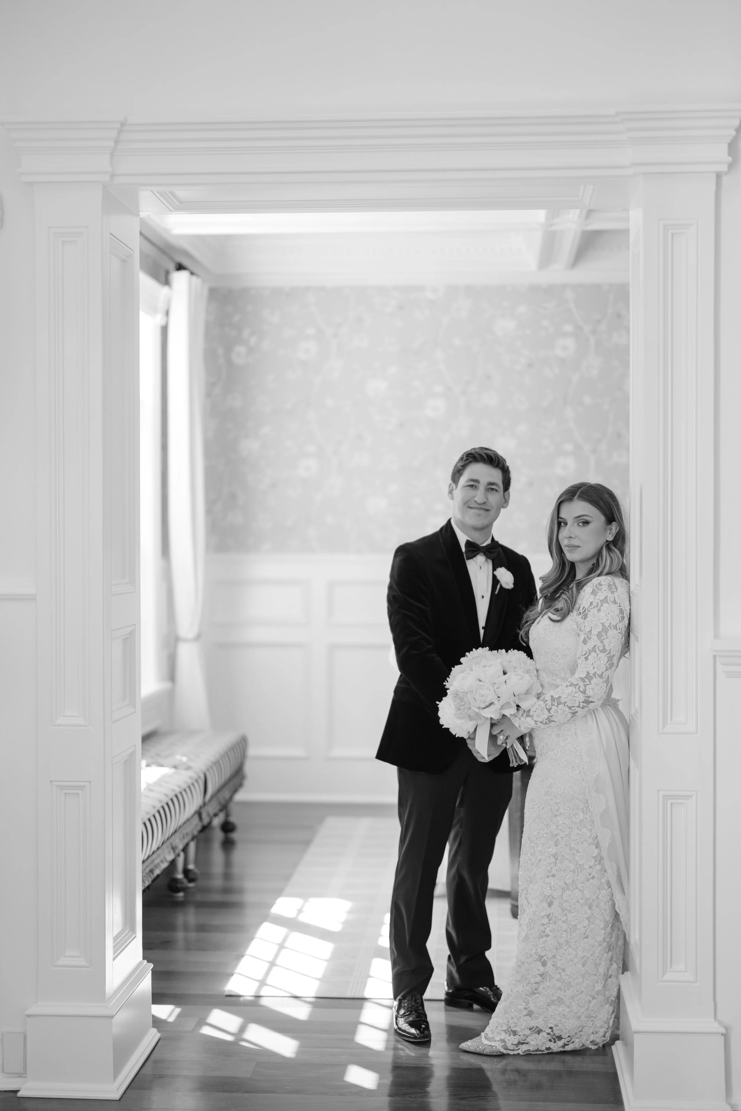 A black-and-white photo of a bride and groom in formal wedding attire standing in a doorway inside a house. The groom is dressed in a tuxedo holding a bouquet, and the bride is in a lace wedding gown holding a bouquet. They are smiling and looking at the camera.
