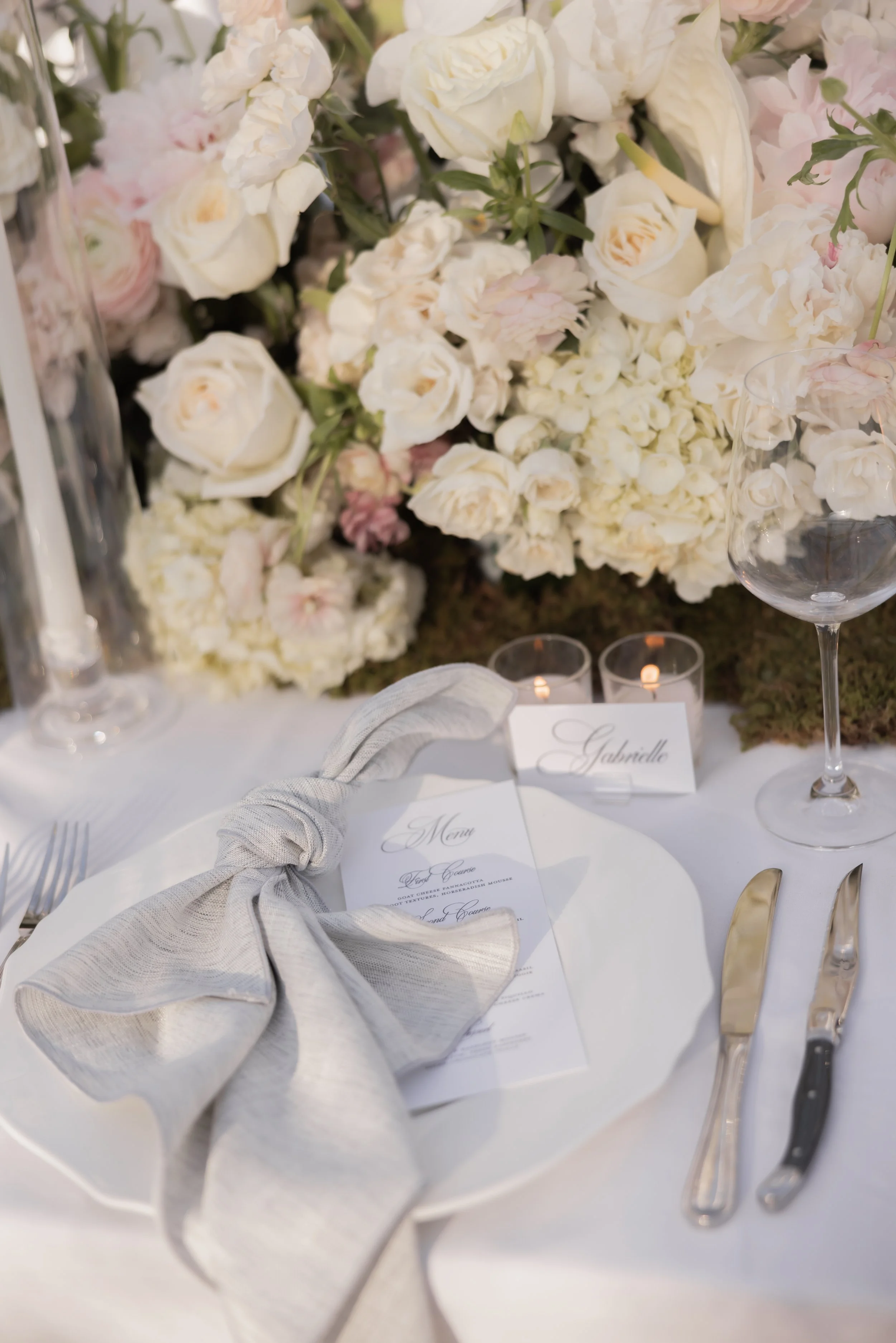 Table setting with a gray cloth napkin tied in a knot, place cards for 'Gabrielle' and 'Menu', cutlery, wine glass, candles, and a floral centerpiece of white and pale pink flowers.