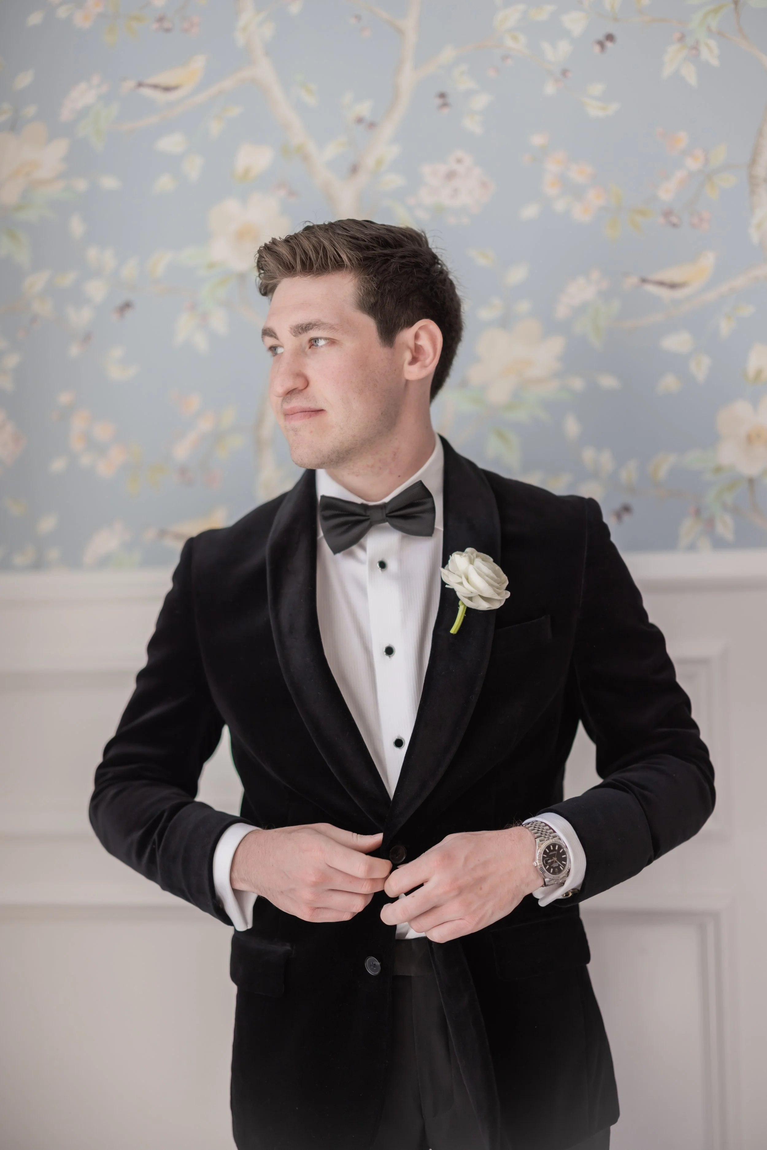 A young man dressed in a black tuxedo with a bow tie and a white boutonniere, sitting indoors in front of a wall with a floral and bird pattern, looking to the side with a serious expression.