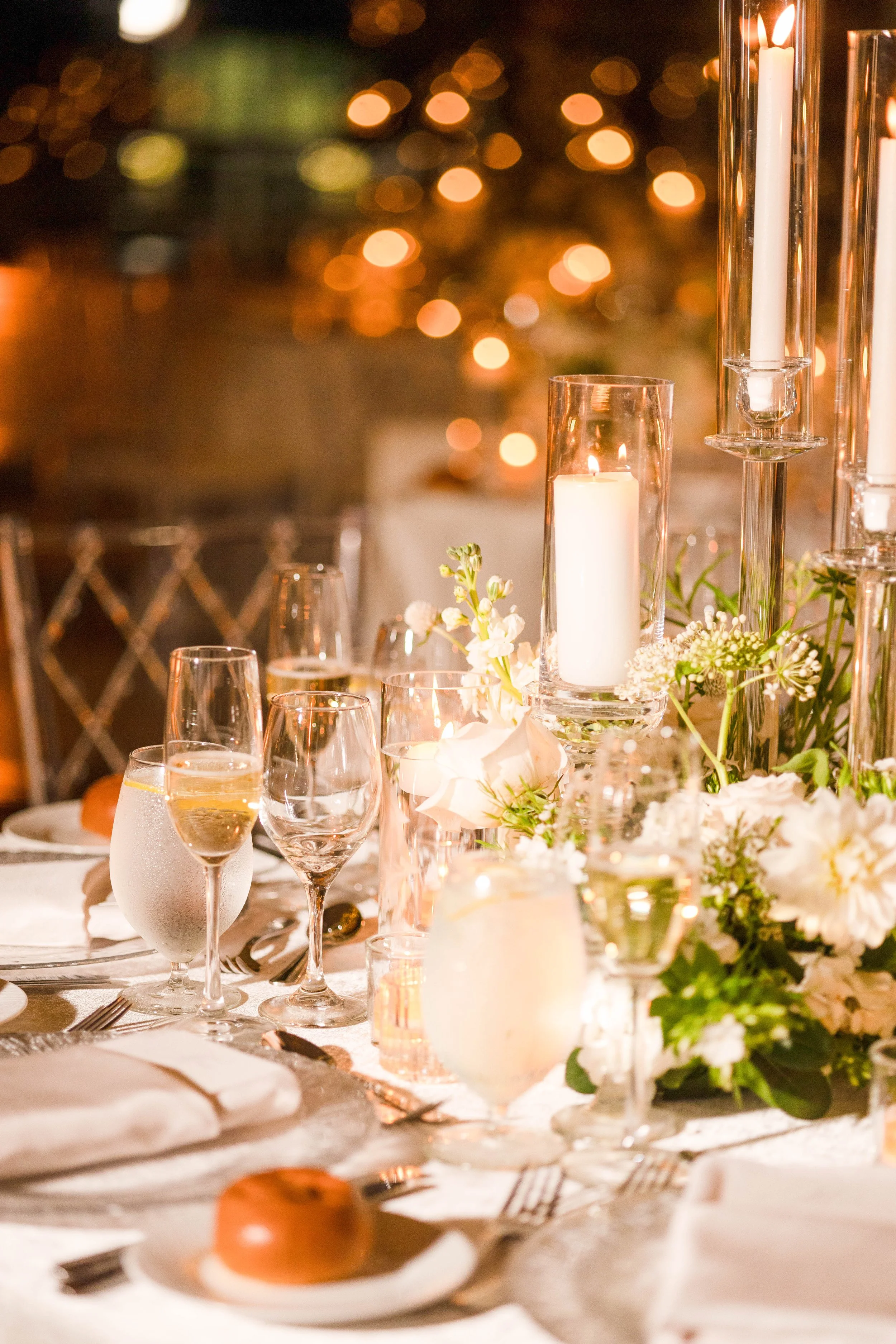 A decorated table with candles, glassware, flowers, and place settings for a formal event or celebration, with bokeh lights in the background.