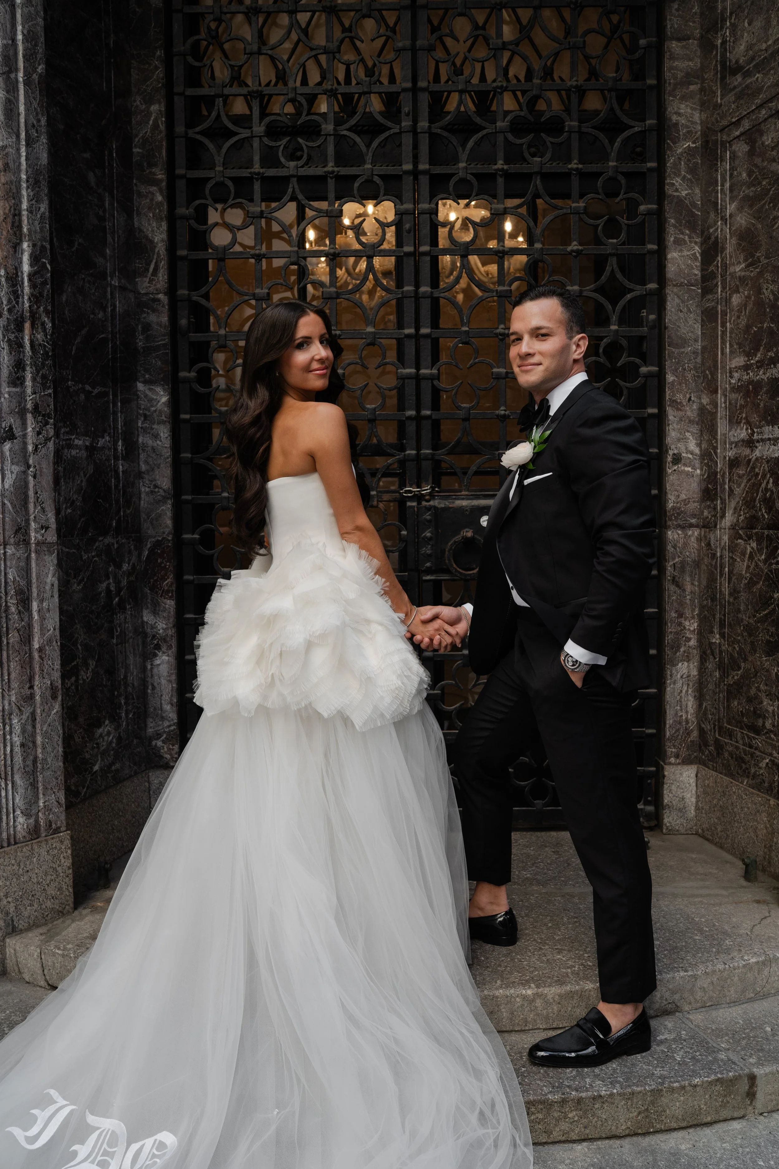 A couple dressed in wedding attire standing outside in front of a decorative metal gate with a chandelier visible behind it. The woman is in a strapless white wedding gown with a full skirt and long, dark wavy hair. The man is in a black tuxedo with bow tie, white shirt, and boutonniere, holding the woman's hand and slightly leaning back with a confident expression.