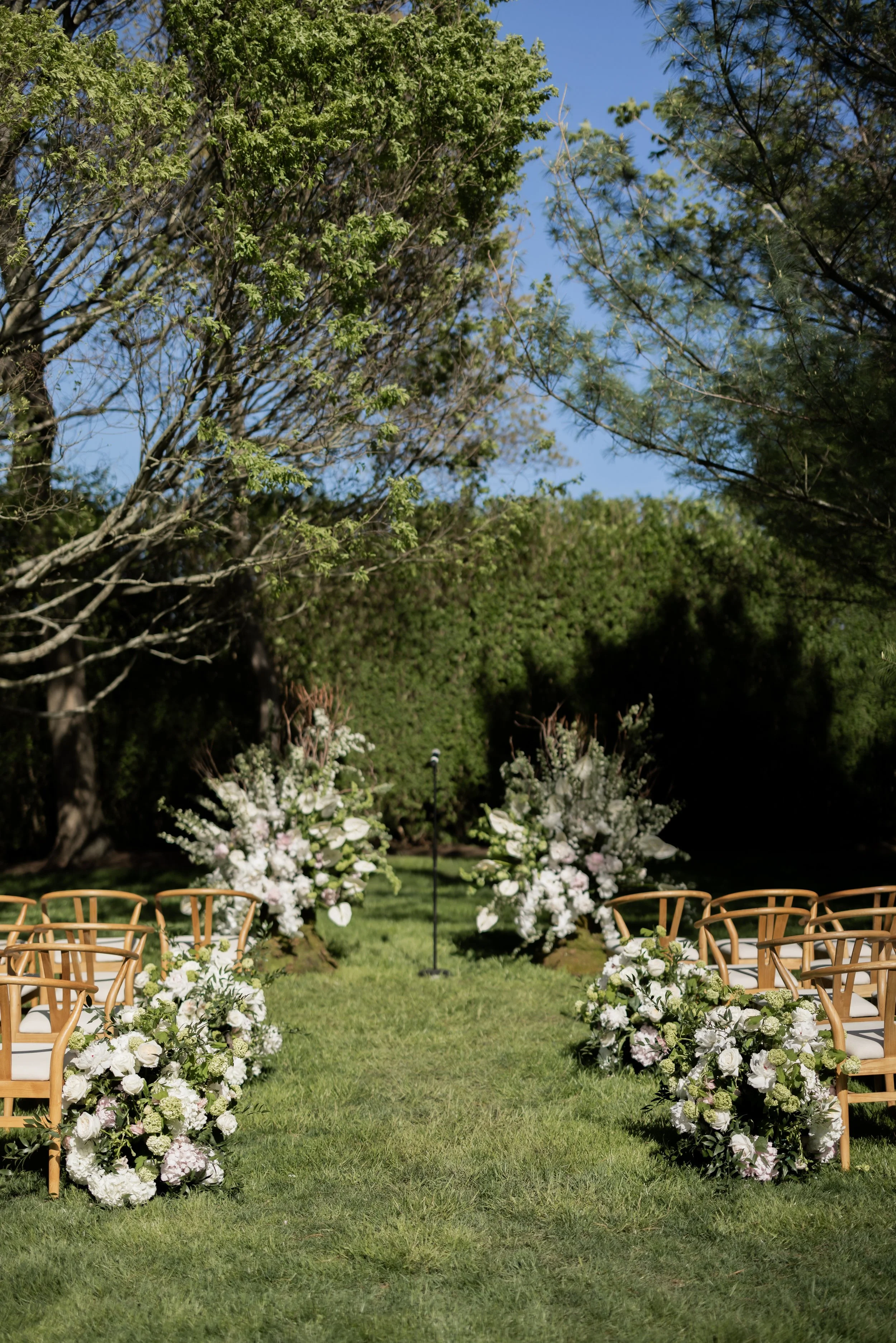 Outdoor wedding setup with an altar decorated with white and pink flowers, arranged along a grassy aisle, surrounded by chairs and tall green trees under a clear blue sky.