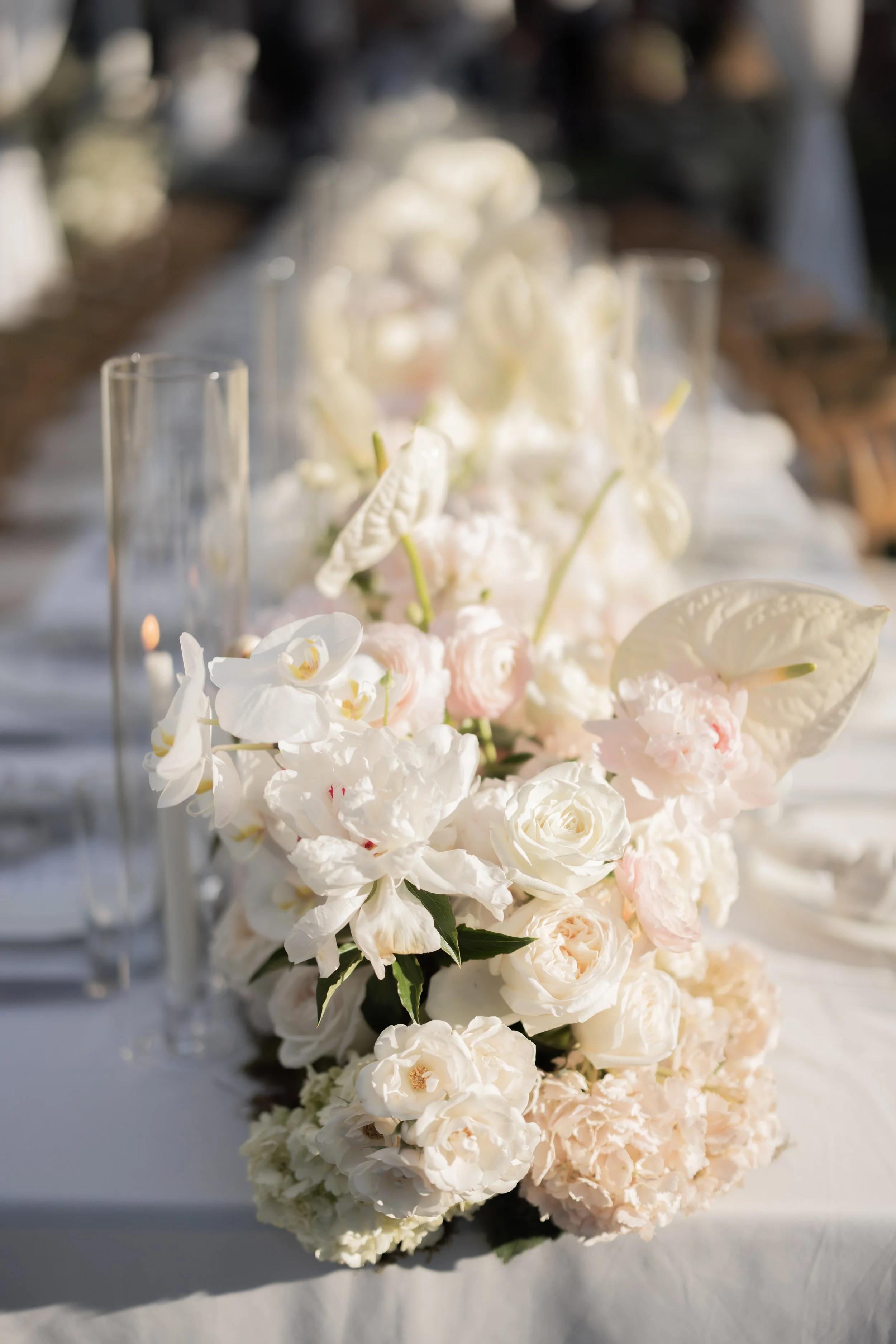 A floral centerpiece with white and pale pink flowers, including roses and orchids, arranged on a table with glassware nearby.