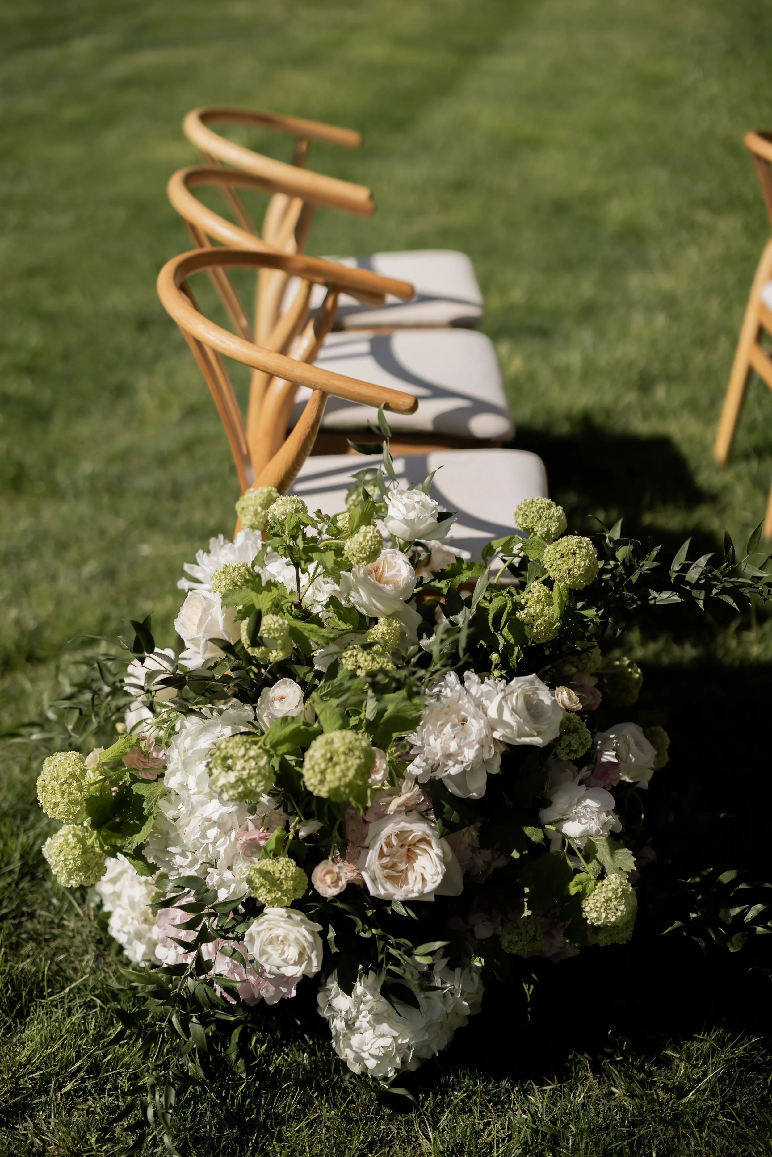 A floral arrangement with white and green flowers and greenery placed outdoors on green grass, with wooden chairs with curved backs in the background.