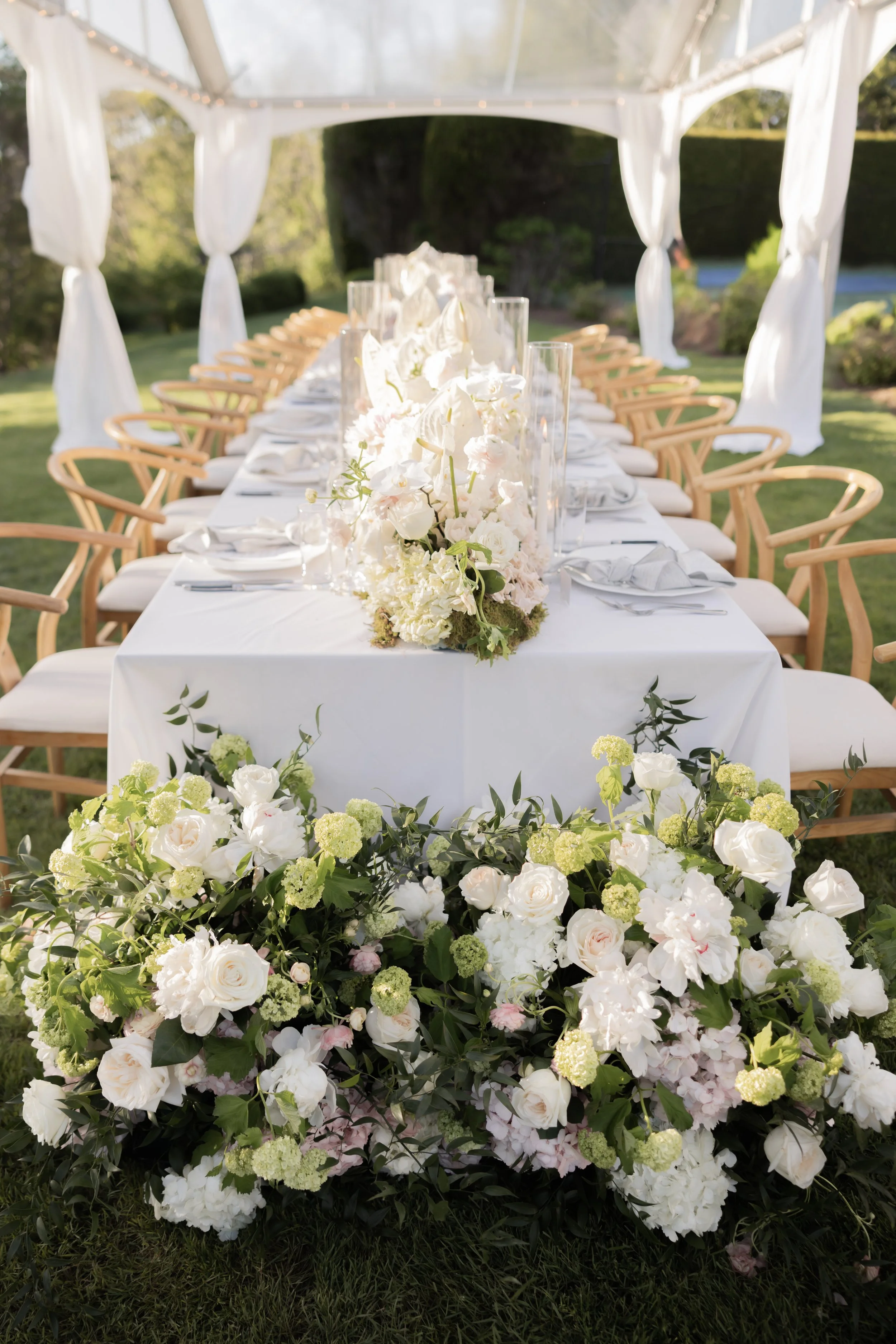 Outdoor wedding reception table adorned with white floral centerpiece and surrounded by chairs, set under a white canopy with draped curtains, with greenery and trees in the background.