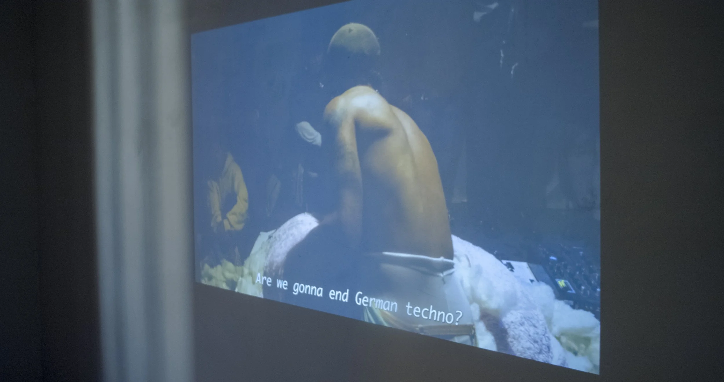 A projection on a wall displaying a person emerging from stuffed animal, with subtitles that read 'Are we gonna end German techno?'
