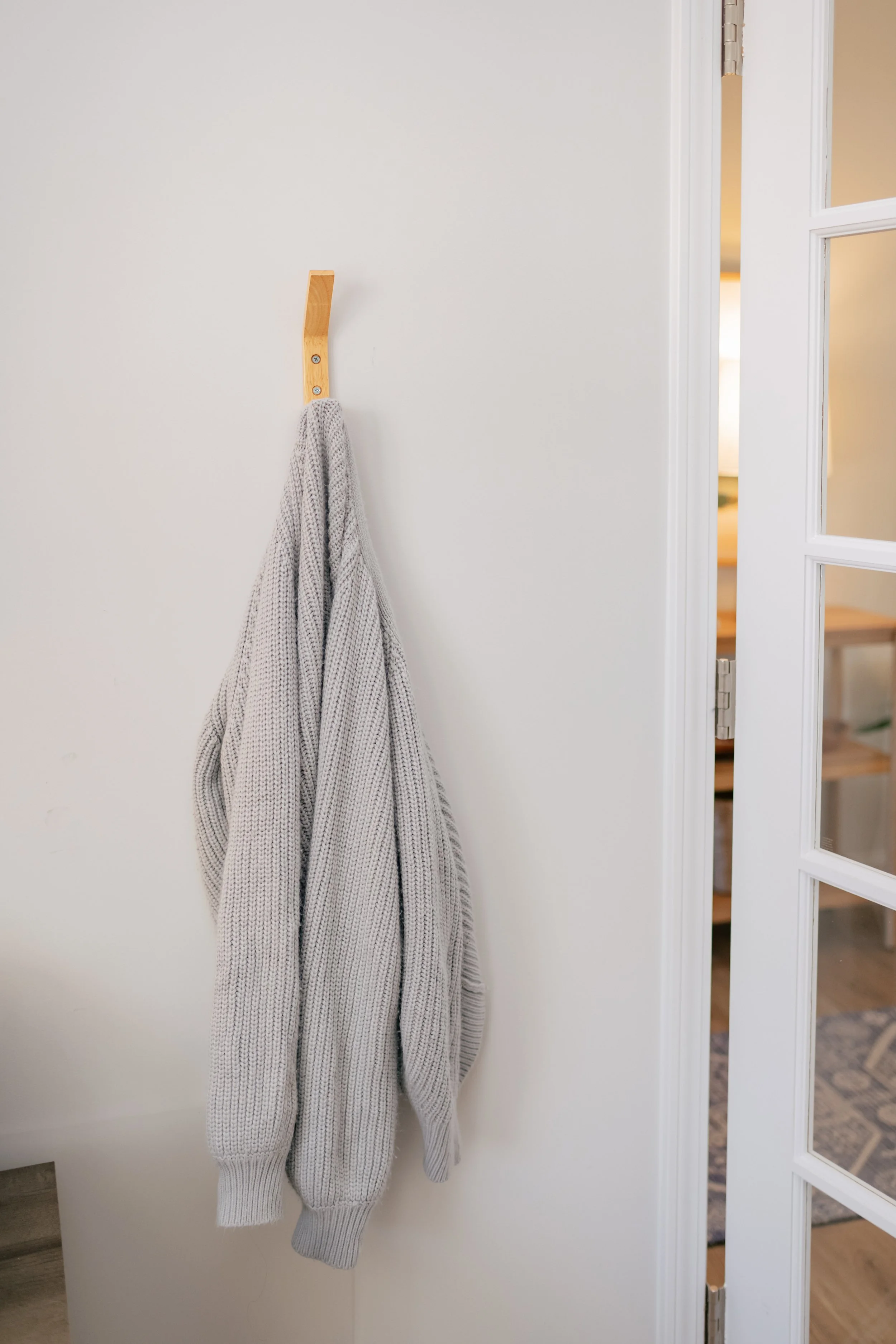 Gray knitted sweater hanging on a wooden hook on a white wall. The sweater has a loose fit and long sleeves.