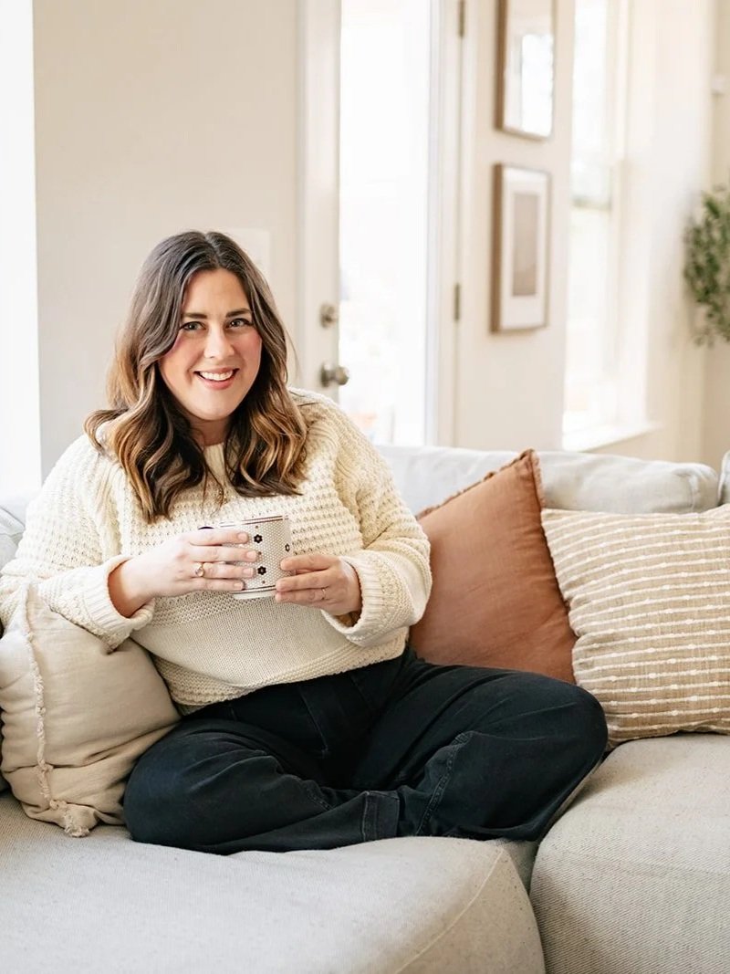 A woman with shoulder-length brown hair smiling while sitting on a beige couch in a brightly lit living room, holding a mug.