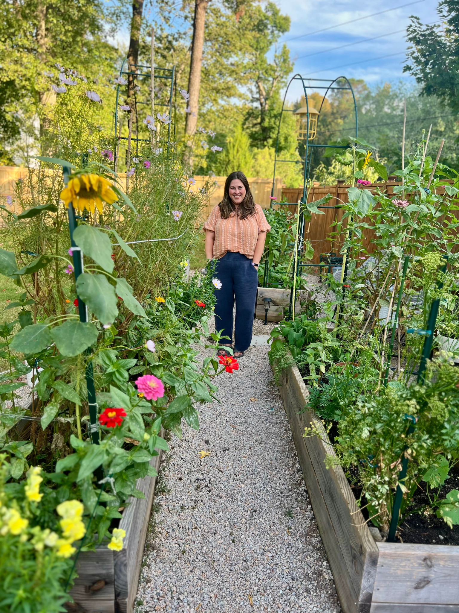 A woman standing on a gravel pathway in a lush garden with raised wooden beds filled with colorful flowers and green plants, surrounded by trees under a blue sky with some clouds.