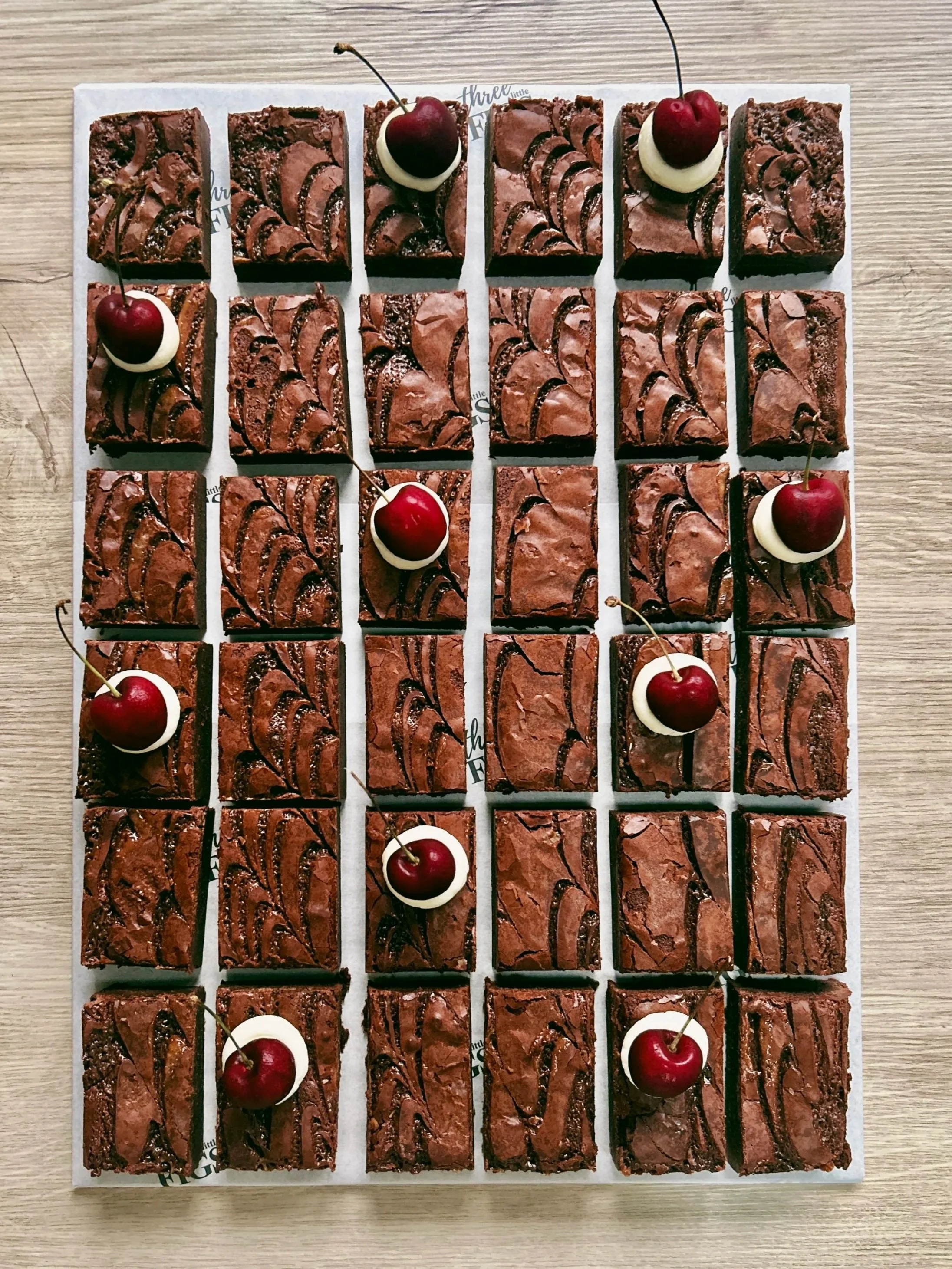 Close-up of rectangular chocolate brownies topped with raspberries and mint leaves, arranged on a white surface outdoors.