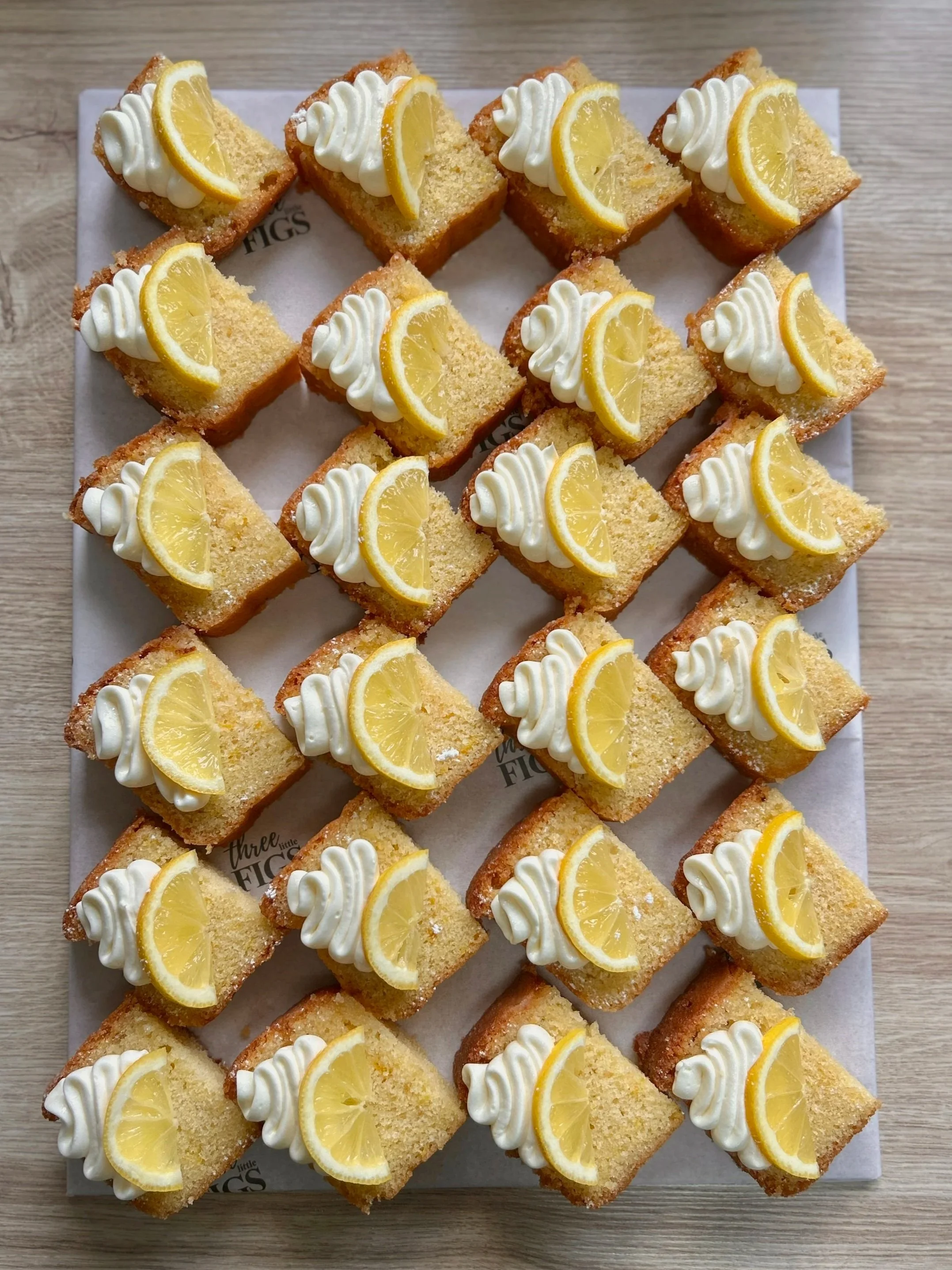 24 small square pieces of lemon drizzle cake topped with white frosting and colorful edible flower petals, arranged on a white tray.