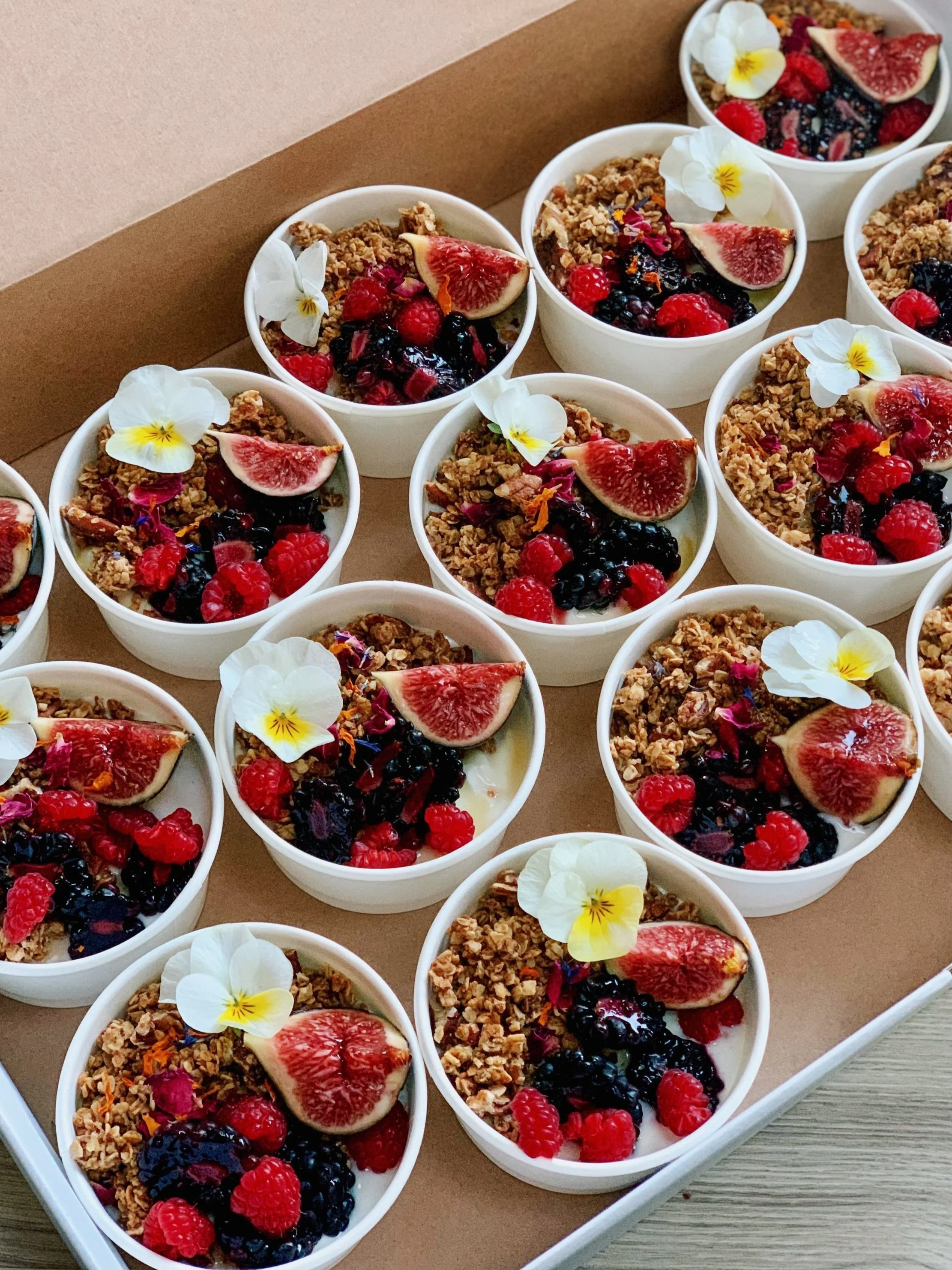 Bowls of yogurt topped with fresh figs, raspberries, blueberries, edible flowers, and granola on a tray.