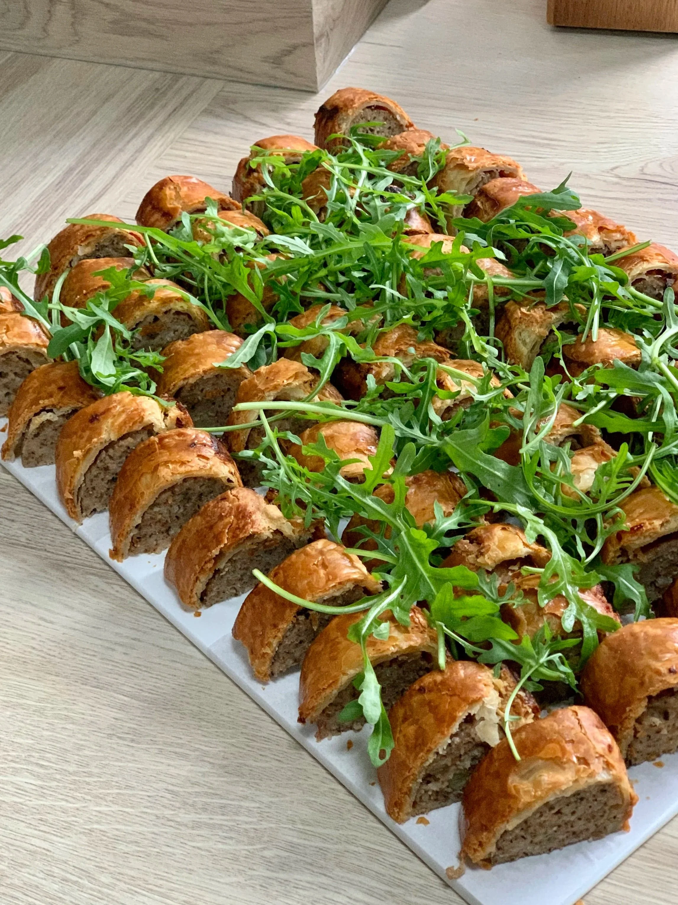 Close-up of a tray of baked sausage rolls garnished with fresh arugula on a wooden surface.