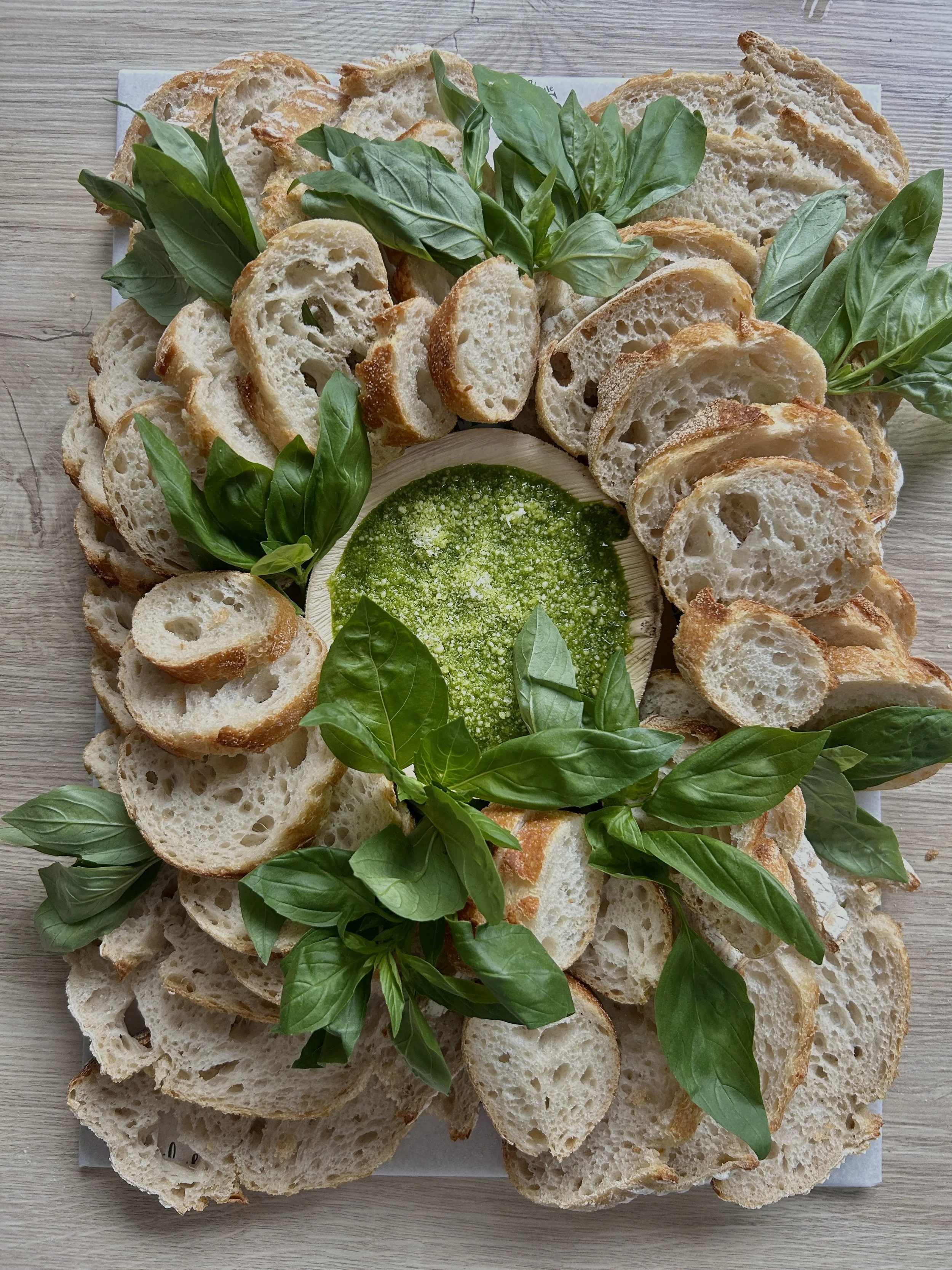 Sliced baguette bread surrounding a bowl of green pesto, garnished with fresh basil leaves and edible flowers.