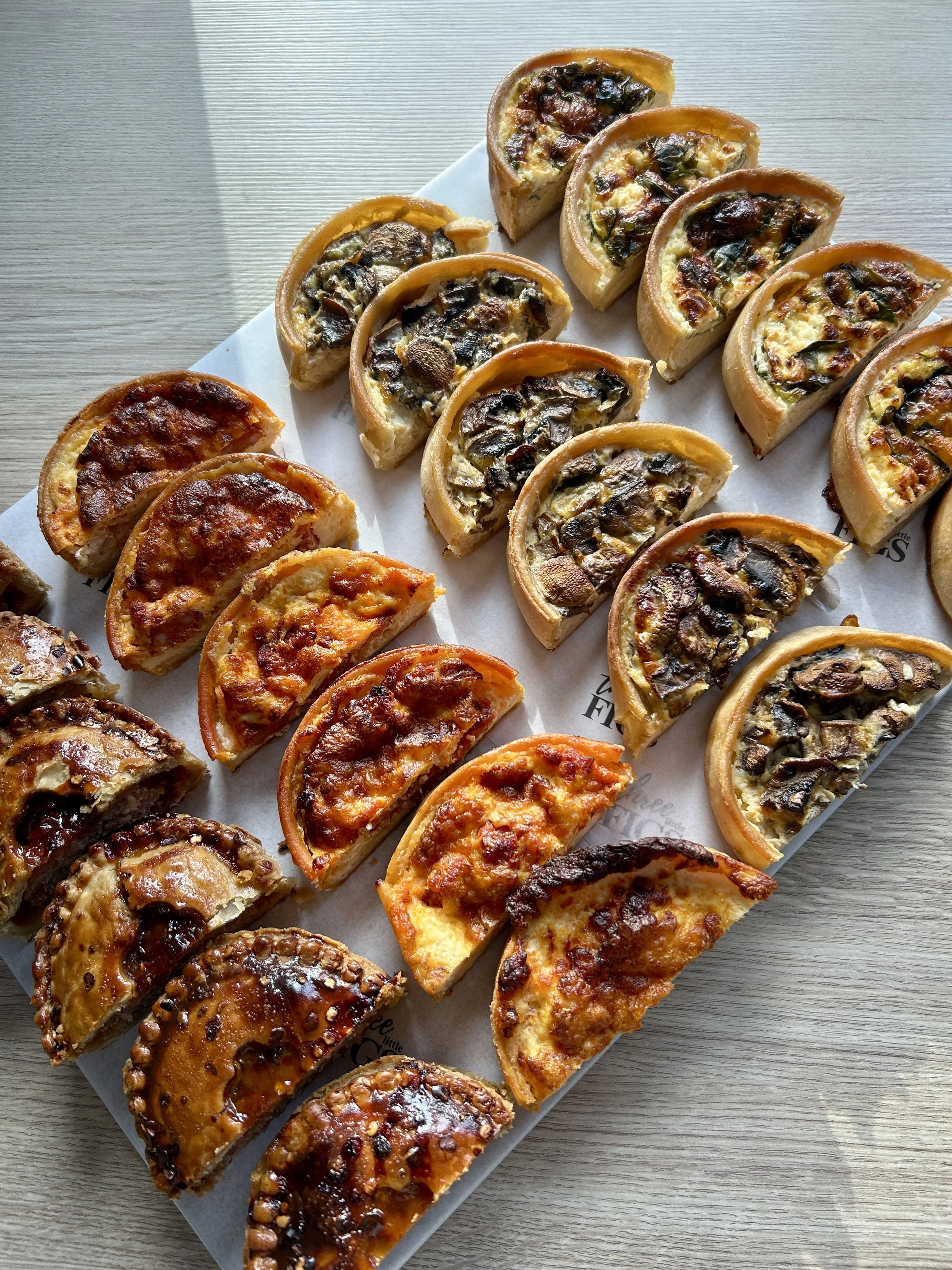 A white rectangular tray with an assortment of mini quiches and savory pastries on a light wooden surface.