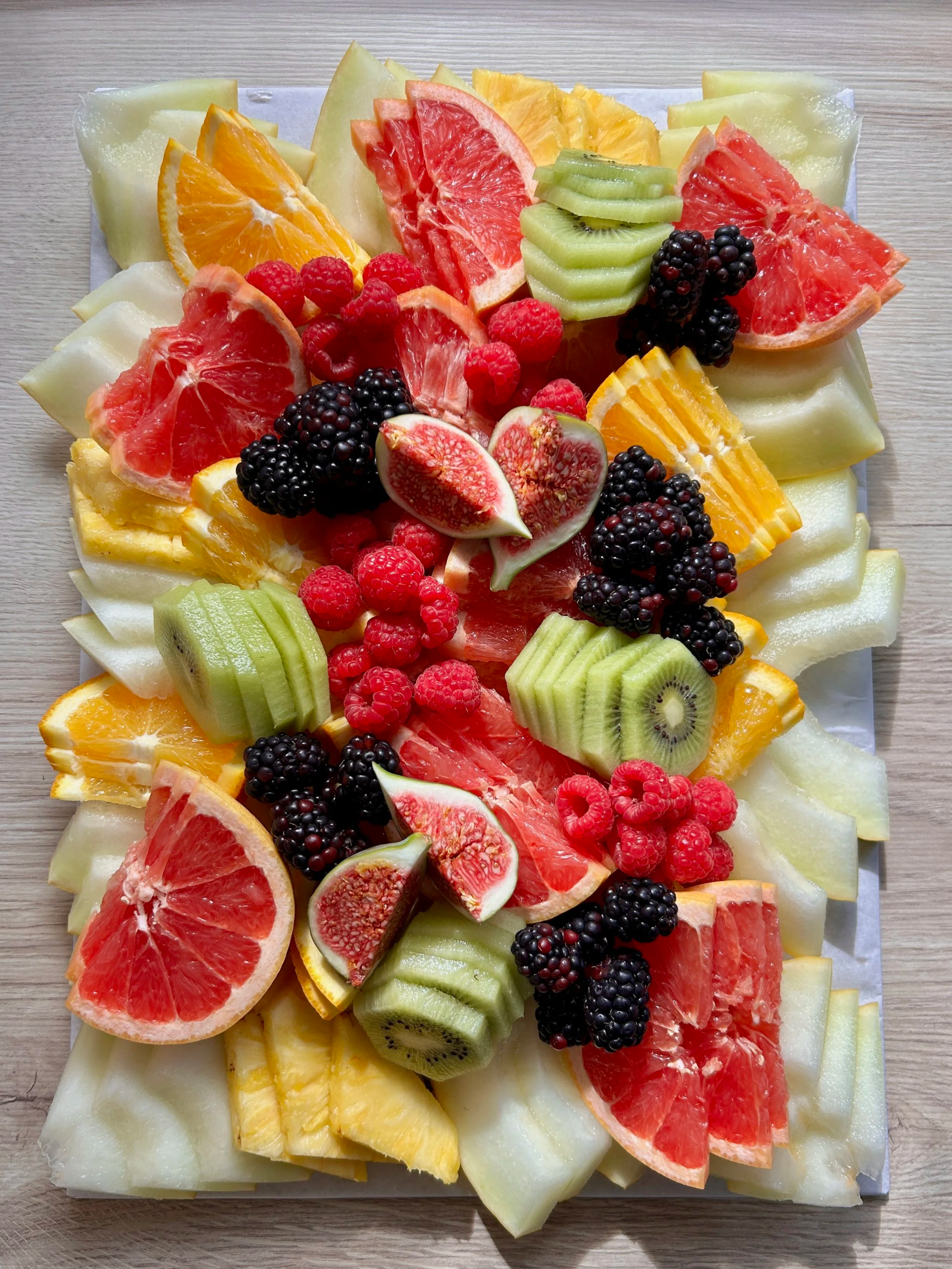 A platter of assorted sliced fruits including watermelon, cantaloupe, honeydew, grapefruit, orange, kiwi, passion fruit, raspberry, blackberry, blueberry, and cherry, garnished with edible flowers.