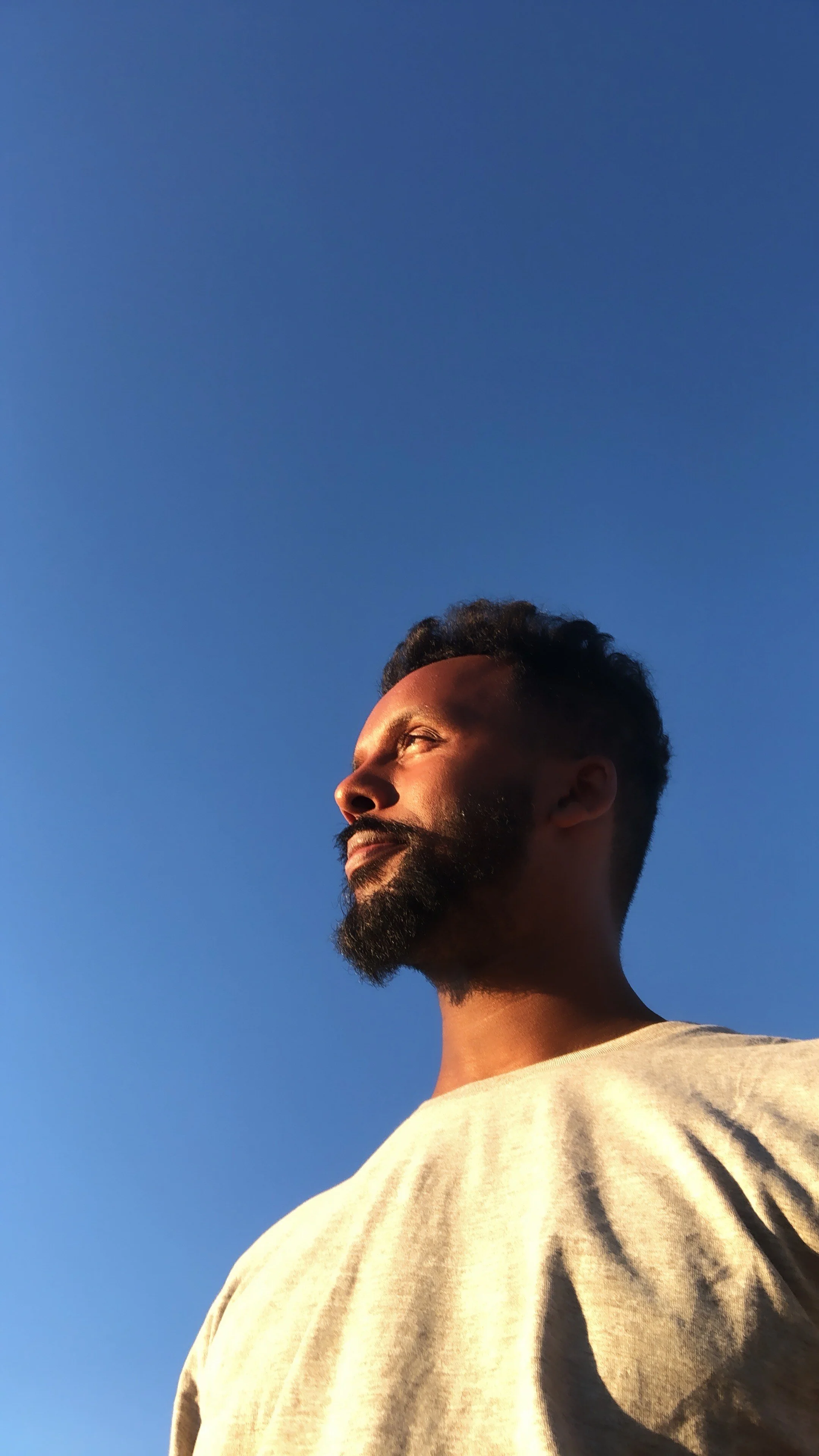 A man with a beard and short curly hair looking towards the sunlight against a clear blue sky.