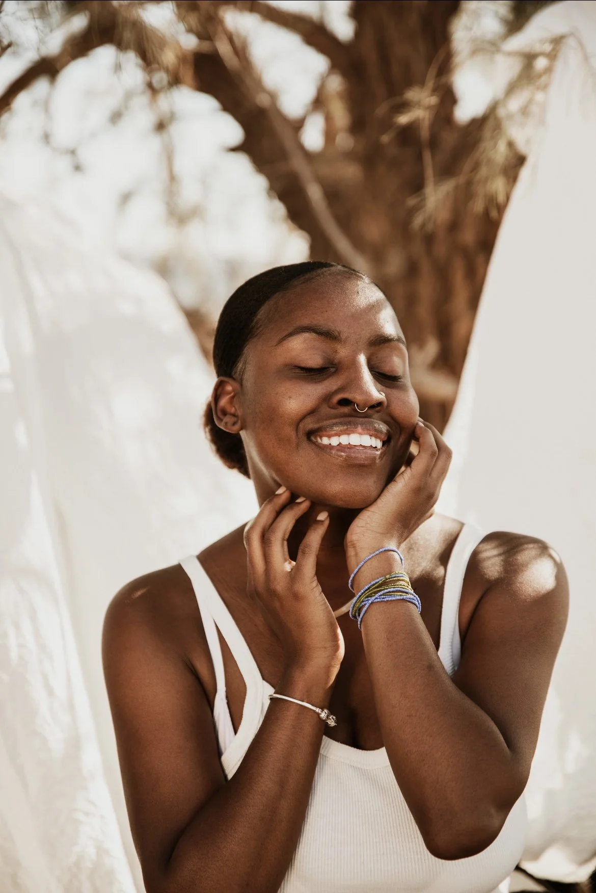 A woman with dark skin, short hair, and a septum piercing is smiling with her eyes closed. She is touching her face gently with both hands. She wears a white tank top and bracelets on her wrists. A large tree with brown branches is in the background.