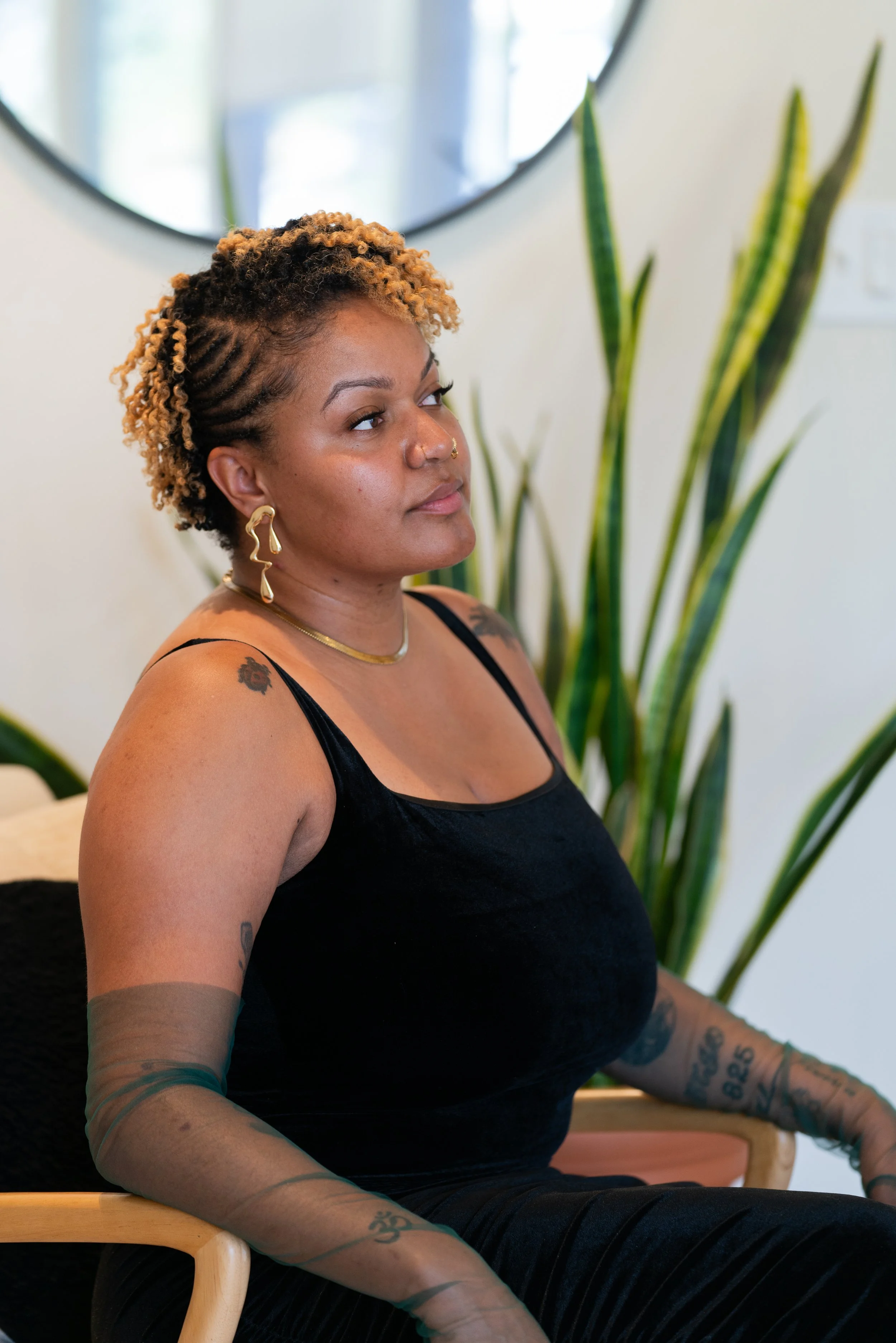 A woman with short, curly, ombré hair and tattoos on her arms, sitting on a chair with a large green plant in the background, wearing a black sleeveless top and gold jewelry.