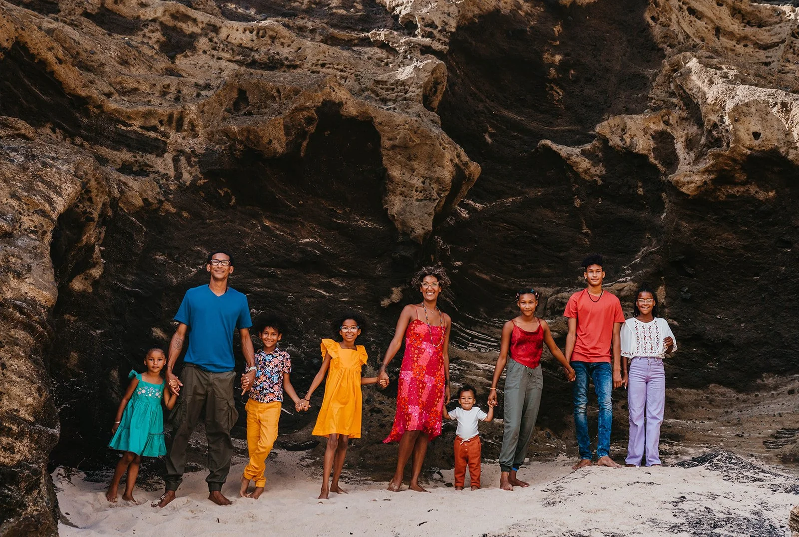 A family of ten people standing hand in hand on a sandy beach with large rock formations behind them, smiling and enjoying the outdoors.