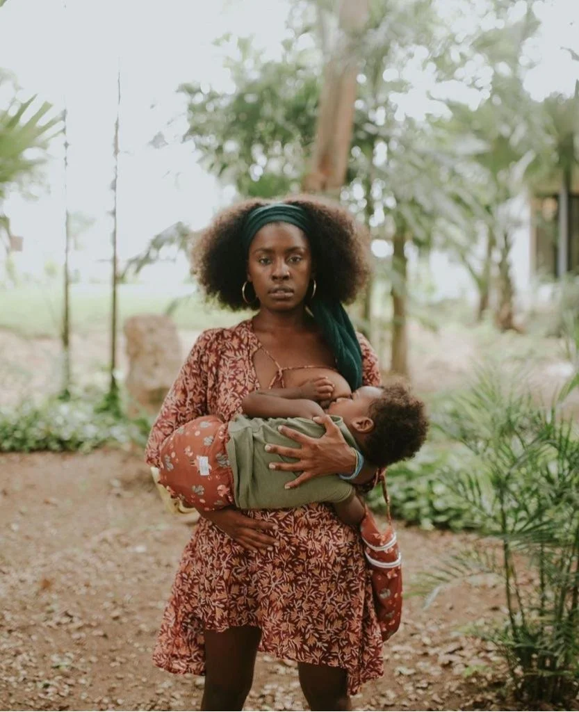 Sumayyah Franklin with curly hair wearing a headscarf and patterned dress holding a young child in her arms outdoors with greenery around.
