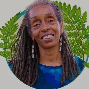 Portrait of a woman with long dreadlocks, wearing earrings, in front of a background with green fern leaves.