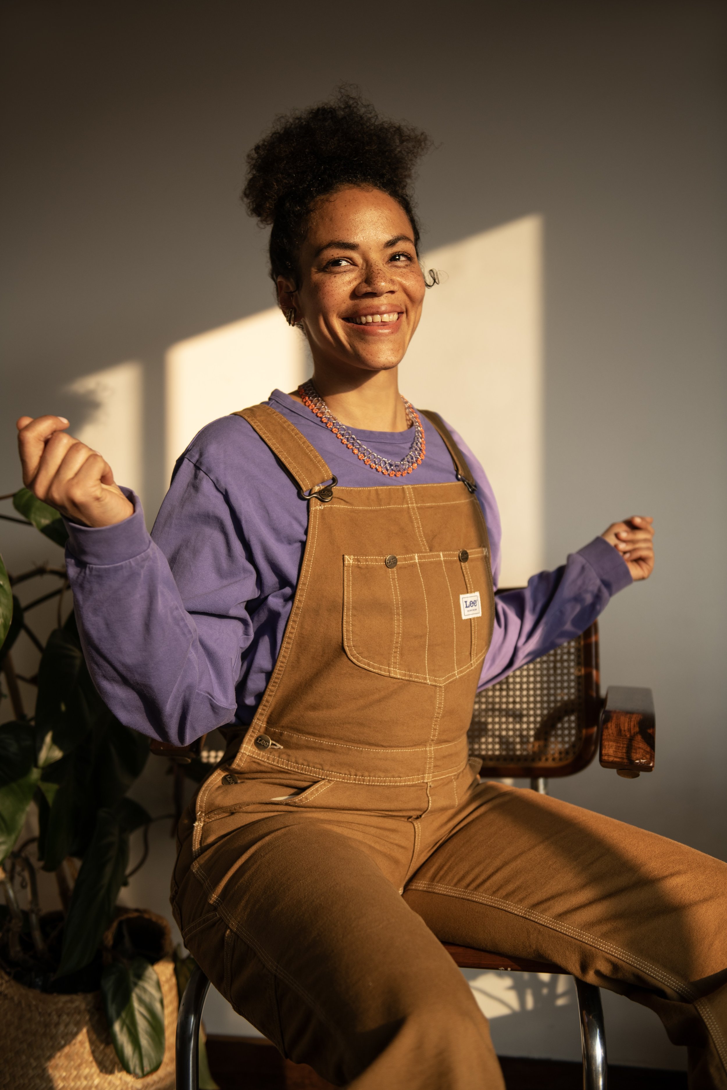 A woman with curly hair, freckles, and a bright smile sitting on a wooden chair, wearing brown overalls over a purple shirt, with a necklace, illuminated by warm sunlight.