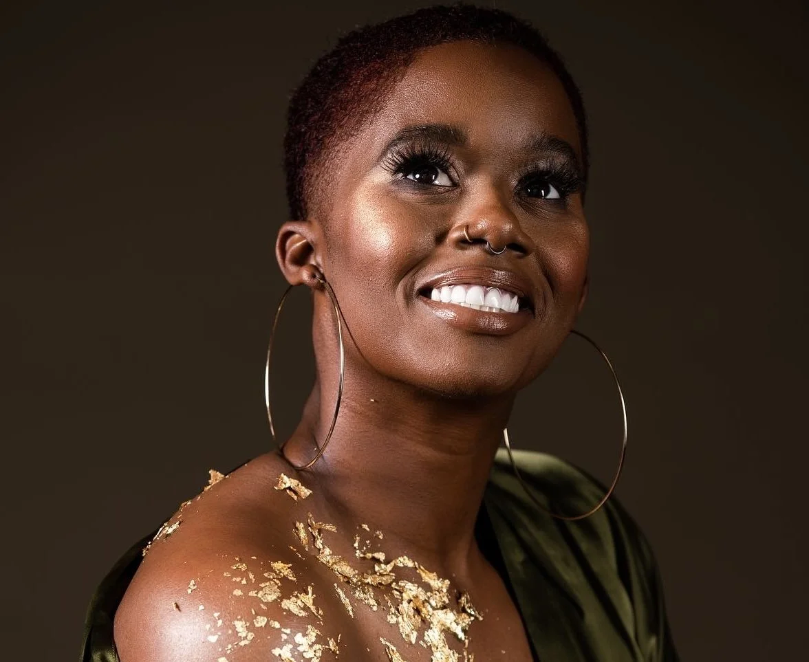 A woman with short red hair, large hoop earrings, a septum piercing, and gold flakes on her shoulder, smiling and looking upward against a dark background.