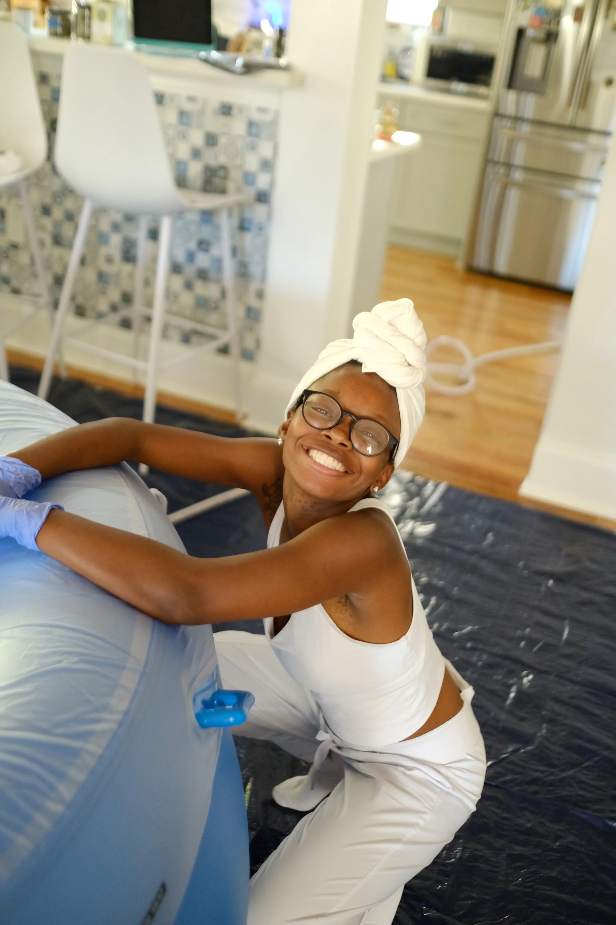 Woman smiling happily, wearing glasses, a white headwrap, white tank top, and white pants, leaning on an air mattress in a kitchen or living room area.