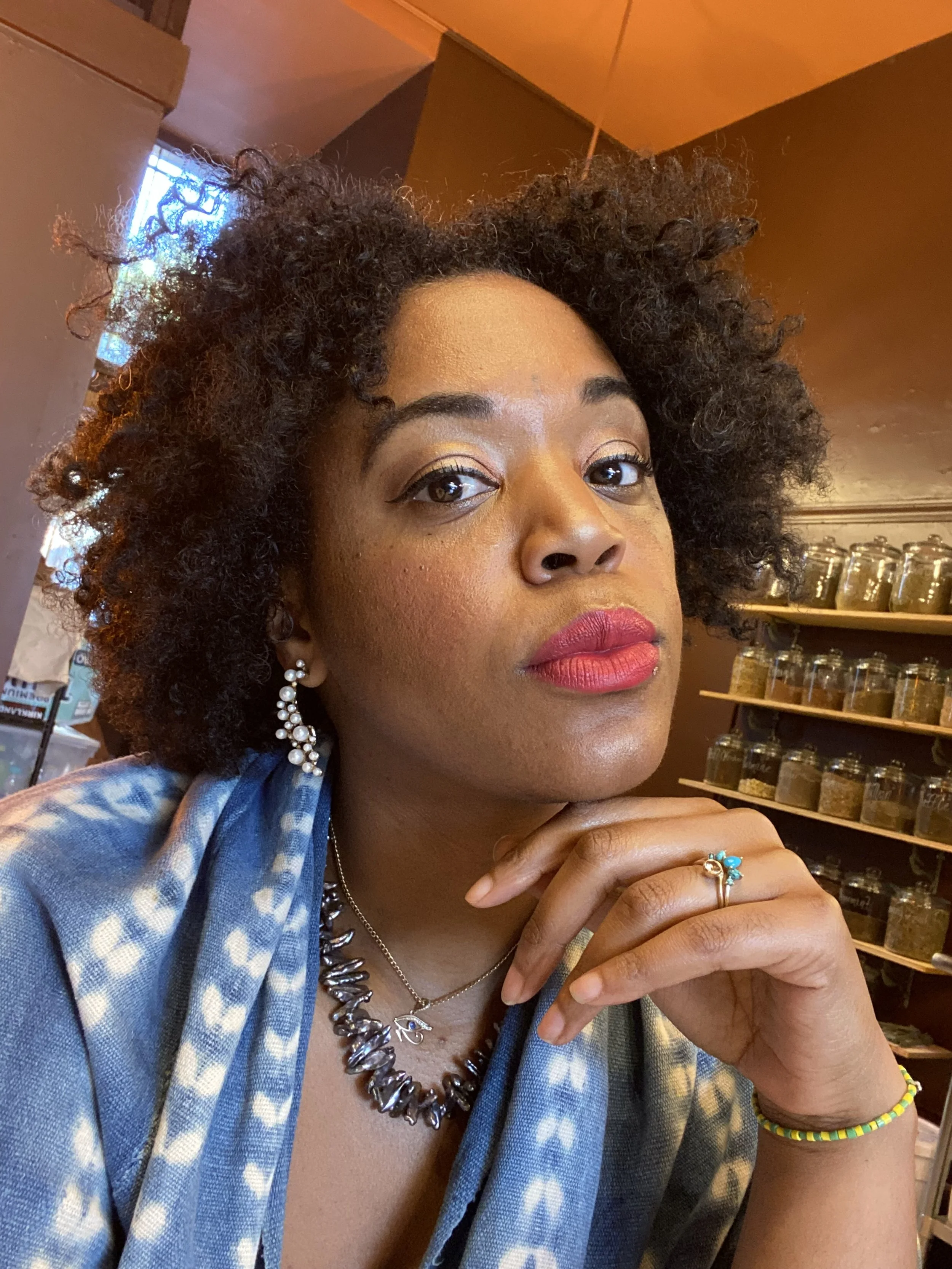 Portrait of a woman with curly hair wearing pearl earrings, a necklace, red lipstick, and a colorful beaded bracelet, sitting in a room with shelves of jars in the background.