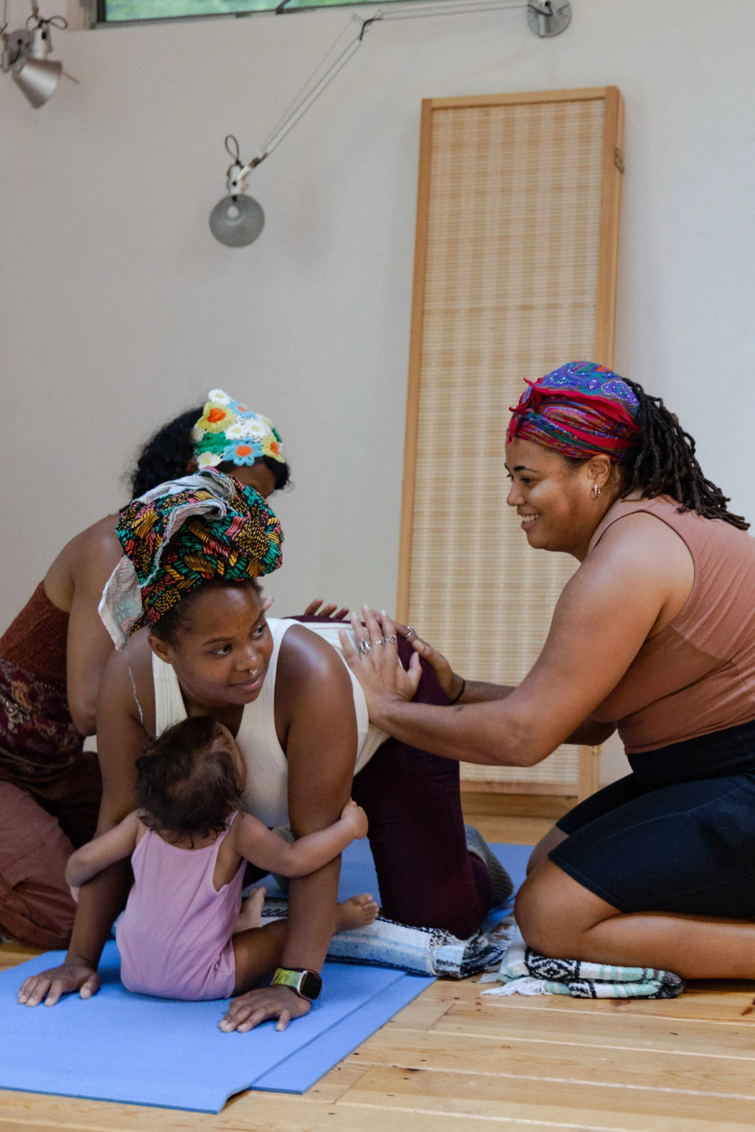 A group of women and a young girl participating in a yoga class, with one woman assisting another and a child hugging her.