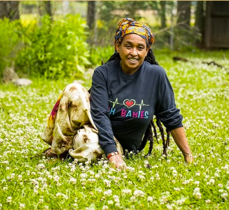 Woman with long braided hair wearing a black sweatshirt with the words 'Oh Babies', sitting on a grassy field with small white flowers, in a garden or yard with trees and a wooden fence in the background.