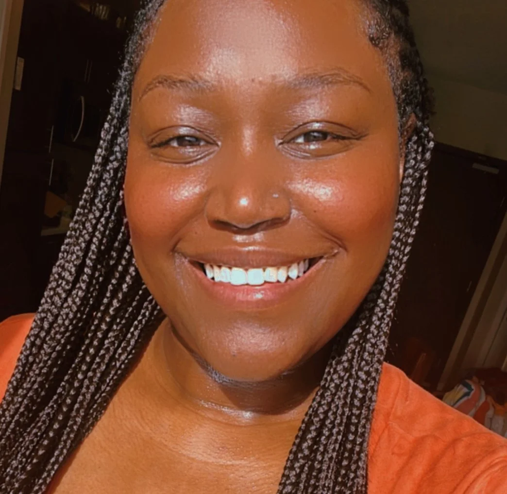 Close-up of a smiling woman with braided hair and wearing an orange top.