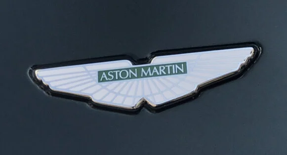 A car emblem with the Aston Martin logo on a dark background.