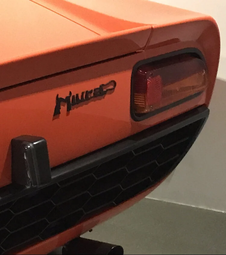 Close-up of the rear end of an orange Lamborghini Miura, showing its taillight, black grille, and Miura badge.