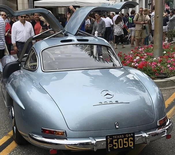 A vintage silver Mercedes-Benz 300SL sports car with gullwing doors open, parked on a city street with a crowd of people and flowers in the background.