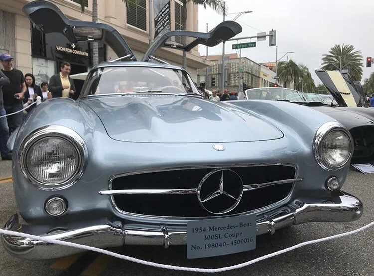 1954 Mercedes-Benz 300SL Coupe displayed at a car show with its gullwing doors open, surrounded by people.