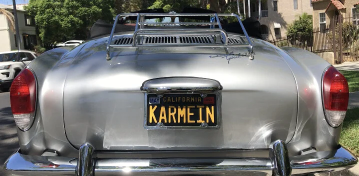 Silver vintage car with California license plate reading 'KARMEIN' parked outdoors, with a luggage rack on top and red taillights.