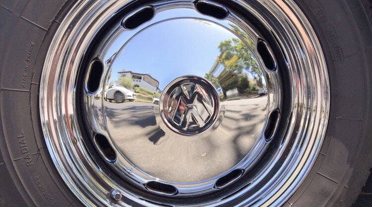 Close-up of a chrome vehicle wheel with VMW logo at the center, reflecting the surrounding street and houses.