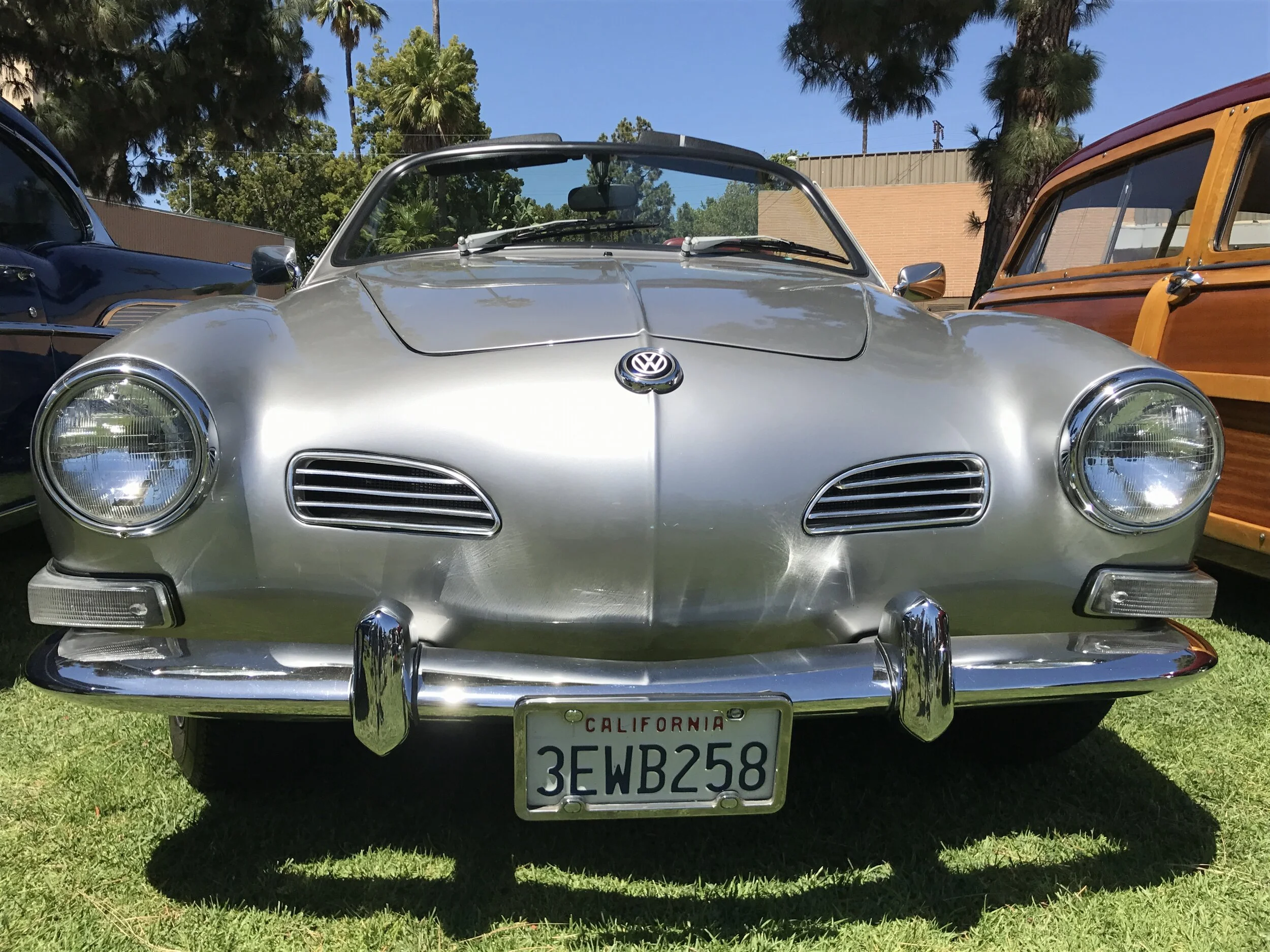 Front view of a vintage silver Volkswagen Karmann Ghia car with California license plate 3EWB258, parked on grass during a car show, surrounded by other classic vehicles, trees, and a building in the background.