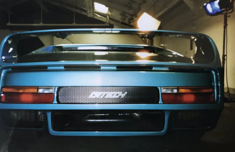 Rear view of a blue race car with a large rear wing and a carbon fiber rear grille in a studio setting with professional lighting equipment.