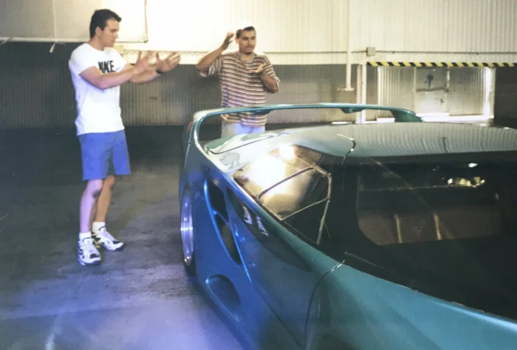 Two men stand near a blue sports car inside a parking garage. One man, wearing a white Nike t-shirt and blue shorts, gestures with his hands. The other man, wearing a striped shirt, is raising one hand near his head and holding a beverage in the othe