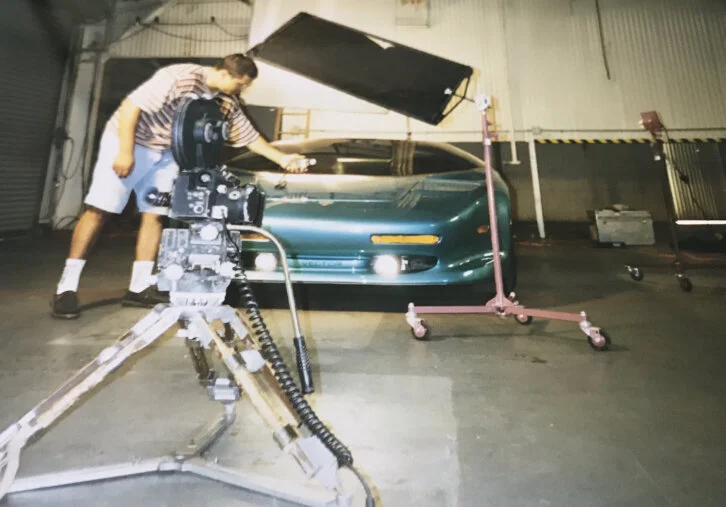 A person is filming a shiny teal sports car in a studio with lighting equipment.
