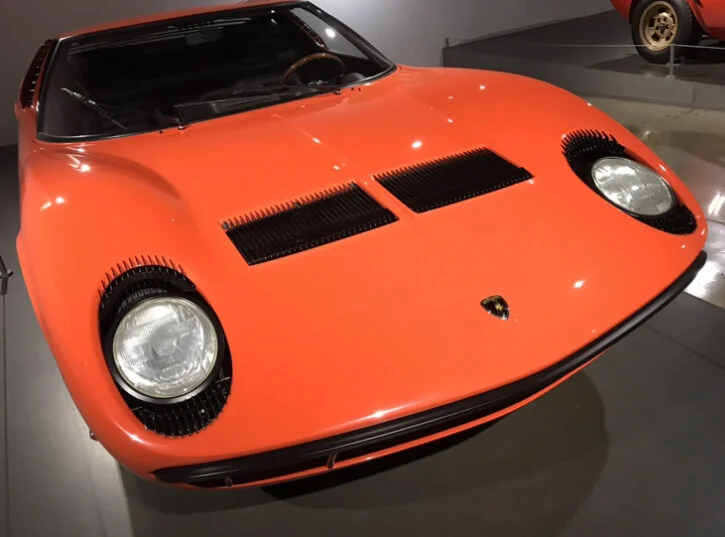 Orange Lamborghini Miura with black vents on hood, round headlights, displayed in a museum.