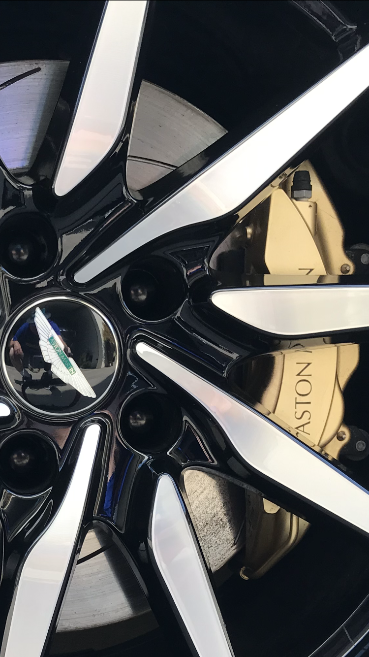 Close-up of a black and silver Aston Martin car wheel with gold brake calipers.
