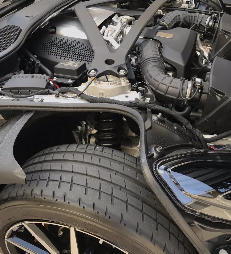 Close-up view of a car engine bay showing the tire, suspension coil spring, and engine components including air intake hose and engine cover.